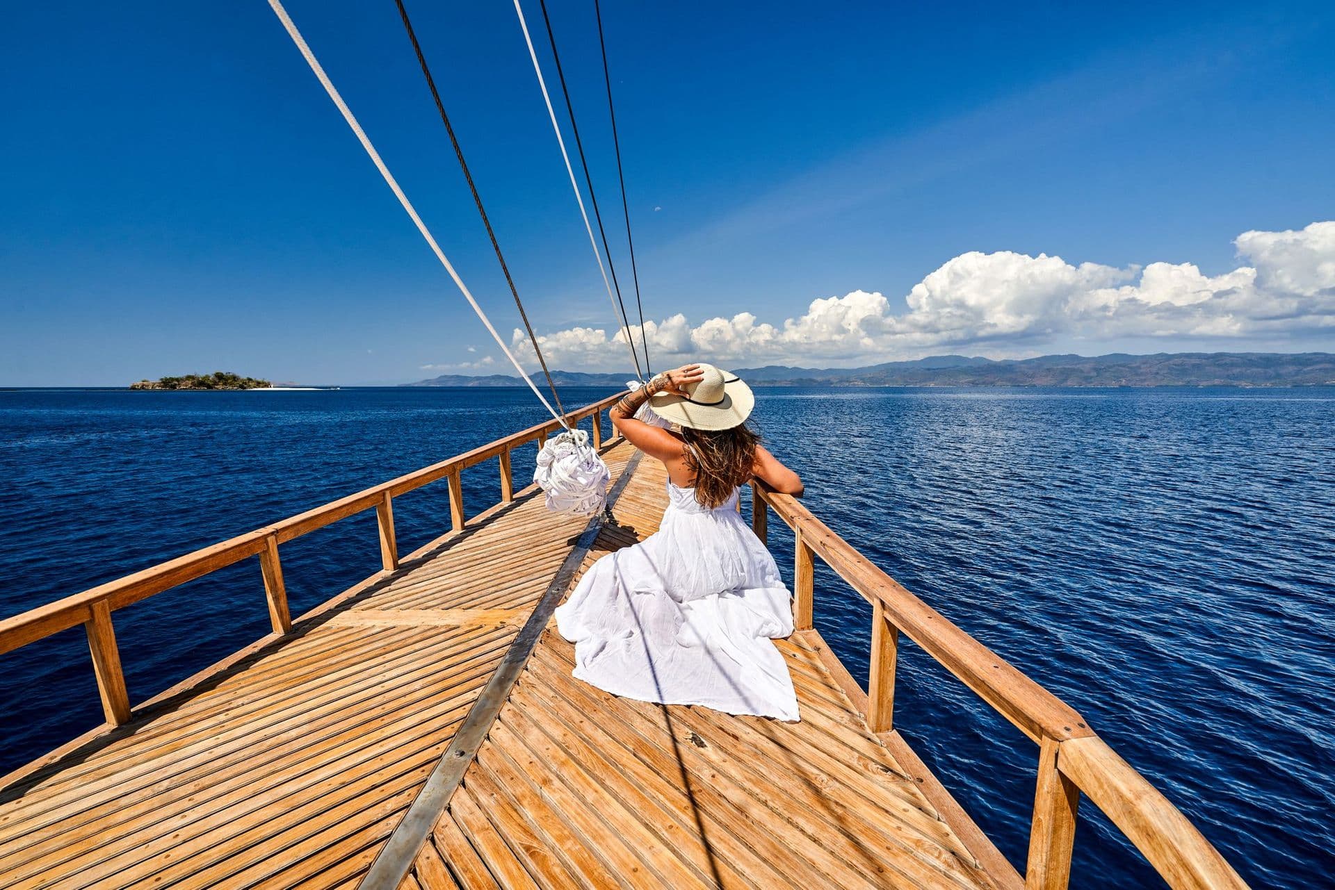 a woman sit on the front deck, sail rope, wooden surface, blue sky, open wide ocean